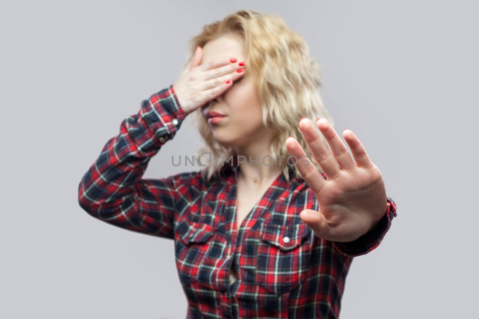 Stop, i dont want to see this. Portrait of beautiful blonde young woman in casual red checkered shirt standing with stop gesture and covering her eyes. studio shot, isolated on grey background.