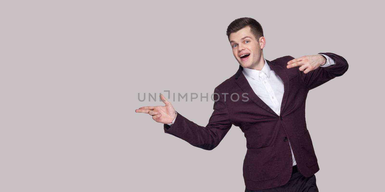 Hey look at this. Portrait of handsome funny young man in suit and white shirt, standing, looking at camera and pointing at copy space with happiness. indoor studio shot, isolated on grey background.