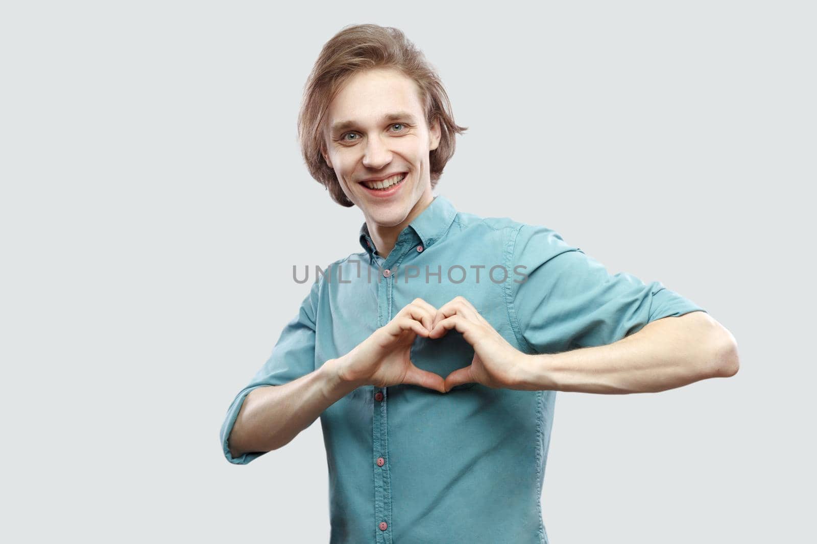 Portrait of happy handsome long haired blonde young man in blue casual shirt standing, toothy smile, love heart gesture hand with toothy smile. indoor studio shot, isolated on light grey background.