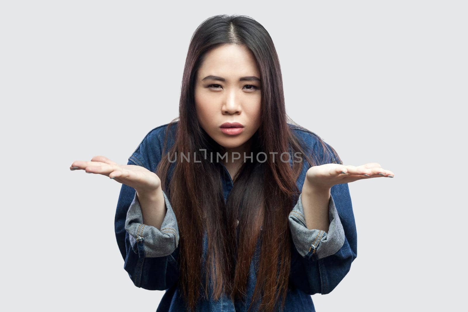 Portrait of anger beautiful brunette asian young woman in casual blue denim jacket with makeup standing raised arms and asking. indoor studio shot, isolated on light grey background.