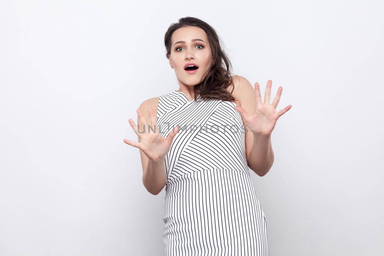 Portrait of scared beautiful young brunette woman with makeup and striped dress standing and looking at camera with blocking hands. indoor studio shot, isolated on grey background.