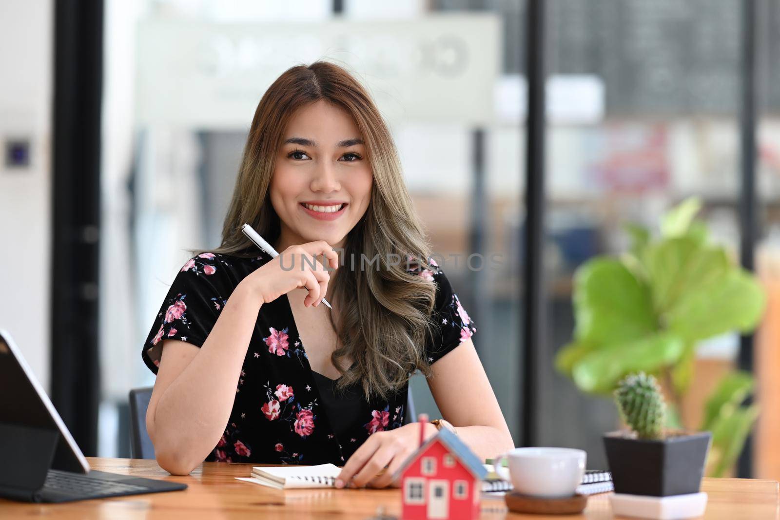 Beautiful real estate agent sitting at her office and smiling to camera. by prathanchorruangsak