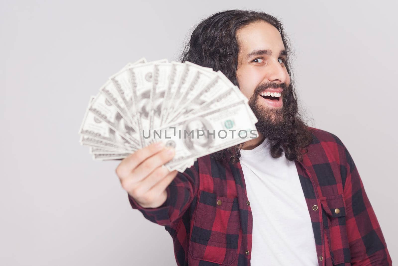 Take it! Happy kidness young businessman in red checkered shirt and long curly hair standing, holding many dollars looking at camera with open mouth. Indoor studio shot, isolated on gray background