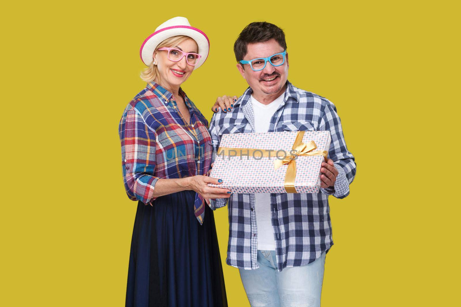 Couple of positive friends, adult man and woman in casual checkered shirt standing beside together holding present gift box, toothy smile and looking. Indoor, isolated, studio shot, yellow background