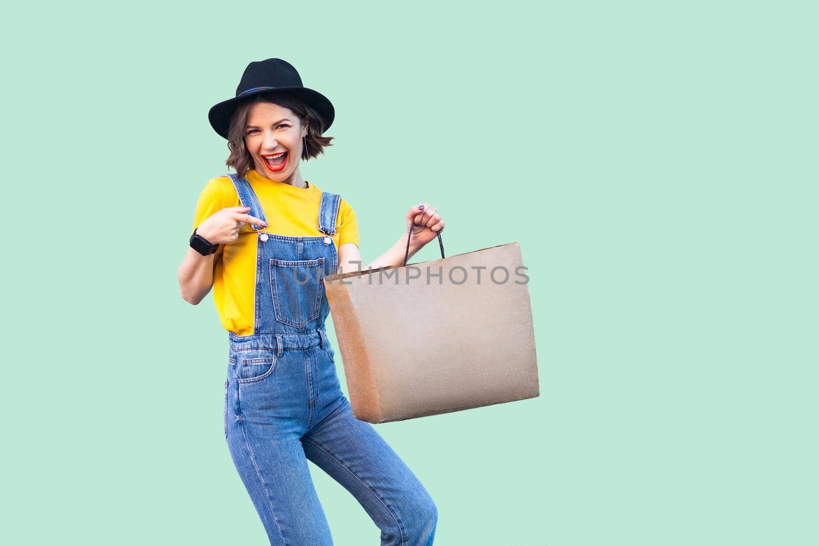 Pleasure beautiful young girl in hipster wear in denim overalls and black hat standing and pointing finger to shopping bag with toothy smile, looking at camera. Studio shot,green background, isolated
