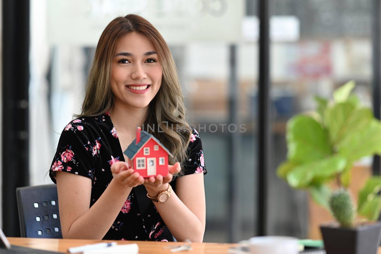 Happy young woman holding small house model and smiling at camera. Real estate investment concept. by prathanchorruangsak