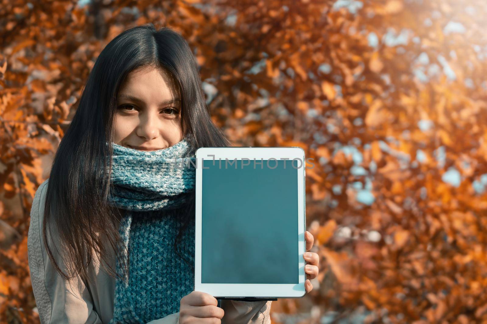 Girl shows the screen of the tablet to the viewer while standing against the backdrop of bright autumn foliage in the park.