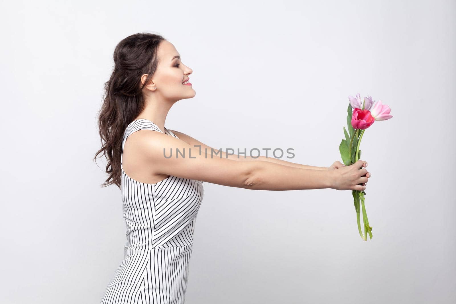 profile side view portrait of young beautiful woman in white striped dress, standing and giving tulips, looking at camera and toothy smiling. indoor studio shot, isolated on light grey background.