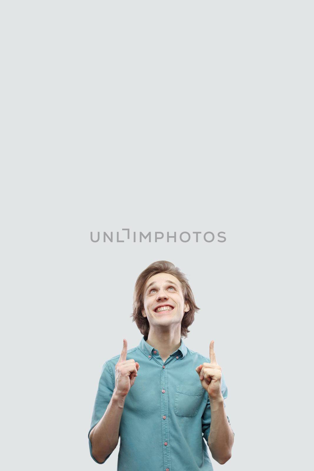 Portrait of happy toothy smiley handsome long haired blonde young man in blue casual shirt standing and pointing at up copyspace. indoor studio shot, isolated on light grey background.