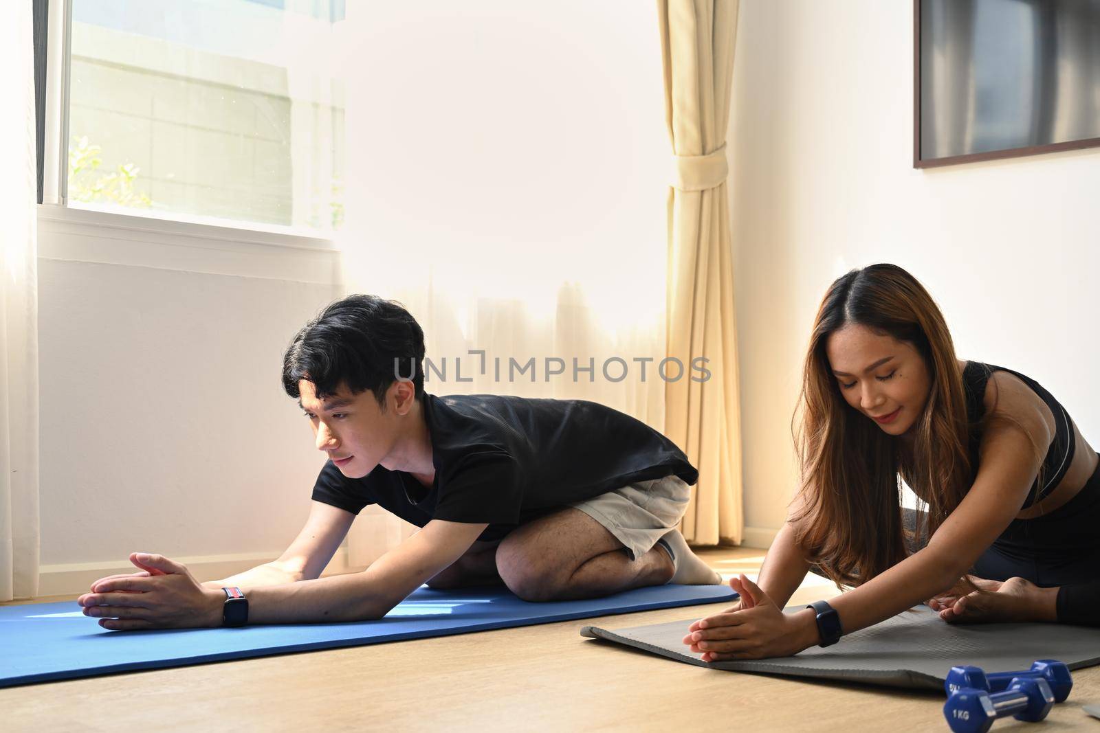 Young asian couple in sports clothes is stretching on yoga mat while working out at home. by prathanchorruangsak