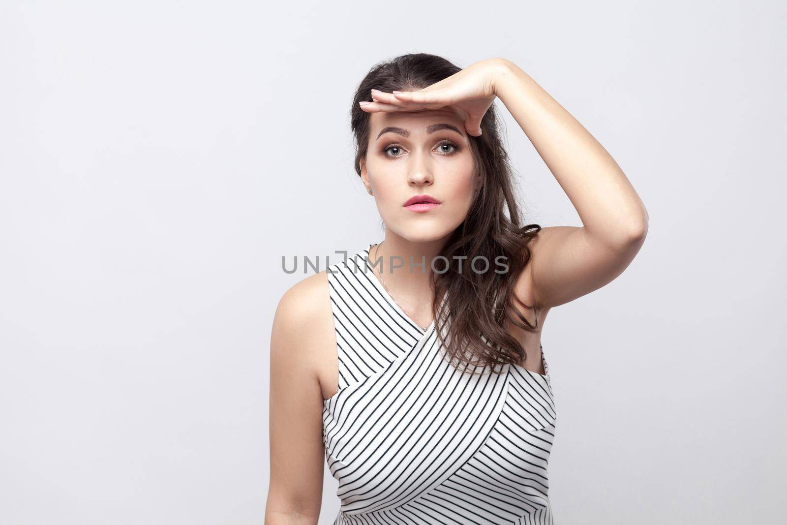 Portrait of beautiful young brunette woman with makeup and striped dress standing, holding hand on head and looking far with serious face. indoor studio shot, isolated on grey background.