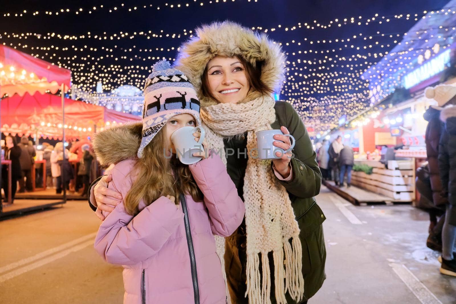 Christmas and New Year holidays, happy mom and daughter kid walking together drinking hot mug tea at Christmas market, sparkling lights of garlands of evening city ferris wheel