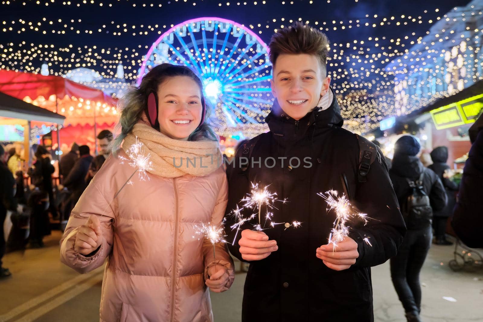 Christmas and New Year holidays, happy teenage couple with sparklers celebrating and having fun at Christmas market, sparkling lights of garlands of evening city ferris wheel background