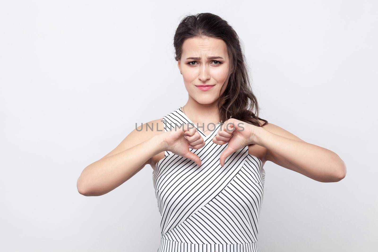 Portrait of sad young brunette woman with makeup and striped dress standing, holding thumbs down and looking at camera with sadness and dislike. indoor studio shot, isolated on grey background.