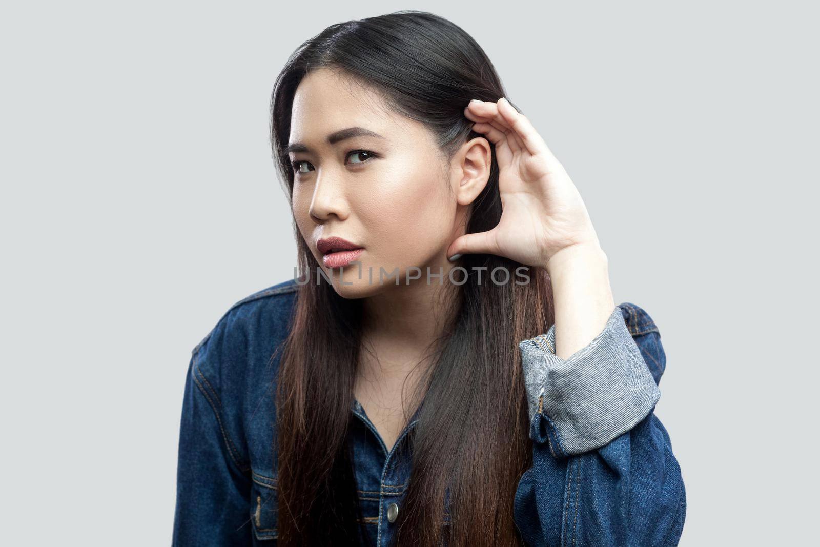 Portrait of attentive beautiful brunette asian young woman in casual blue jacket with makeup standing holding hand near ear and trying to spy or listen. studio shot, isolated on light grey background.
