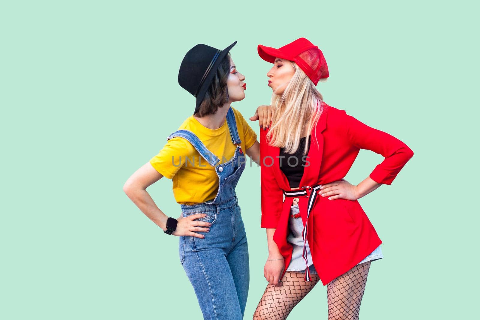 Side view of couple of beautiful stilysh hipster best friends in fashionable clothes standing, posing and sending air kiss, glad to see each other. Indoor studio shot, isolated on green background