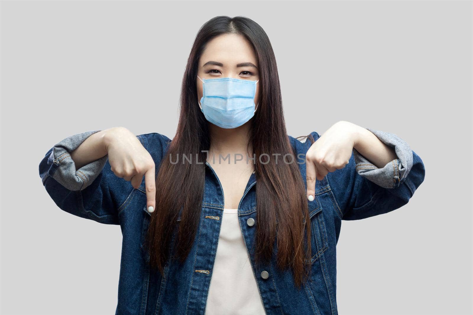 Here and right now. Portrait of serious bossy brunette asian young woman with surgical medical mask in blue denim jacket standing and pointing down. studio shot, isolated on grey background.