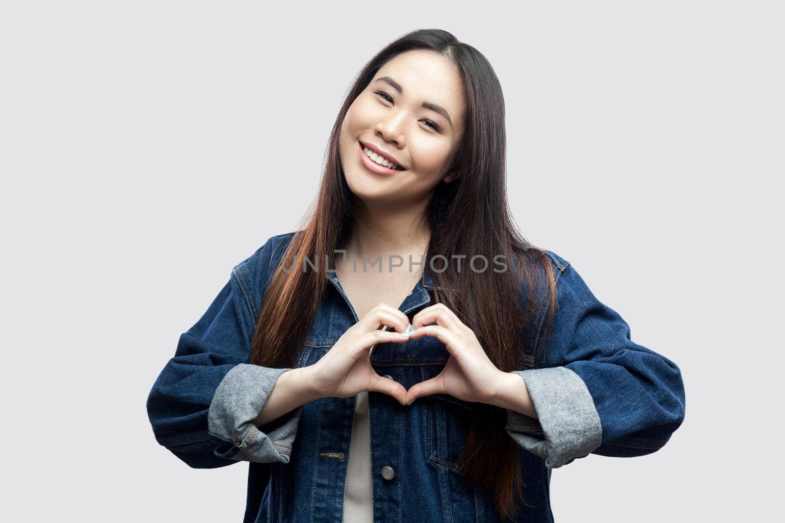 Portrait of happy beautiful brunette asian young woman in casual blue denim jacket with makeup standing with love heart gesture with toothy smile. studio shot, indoor isolated on light grey background