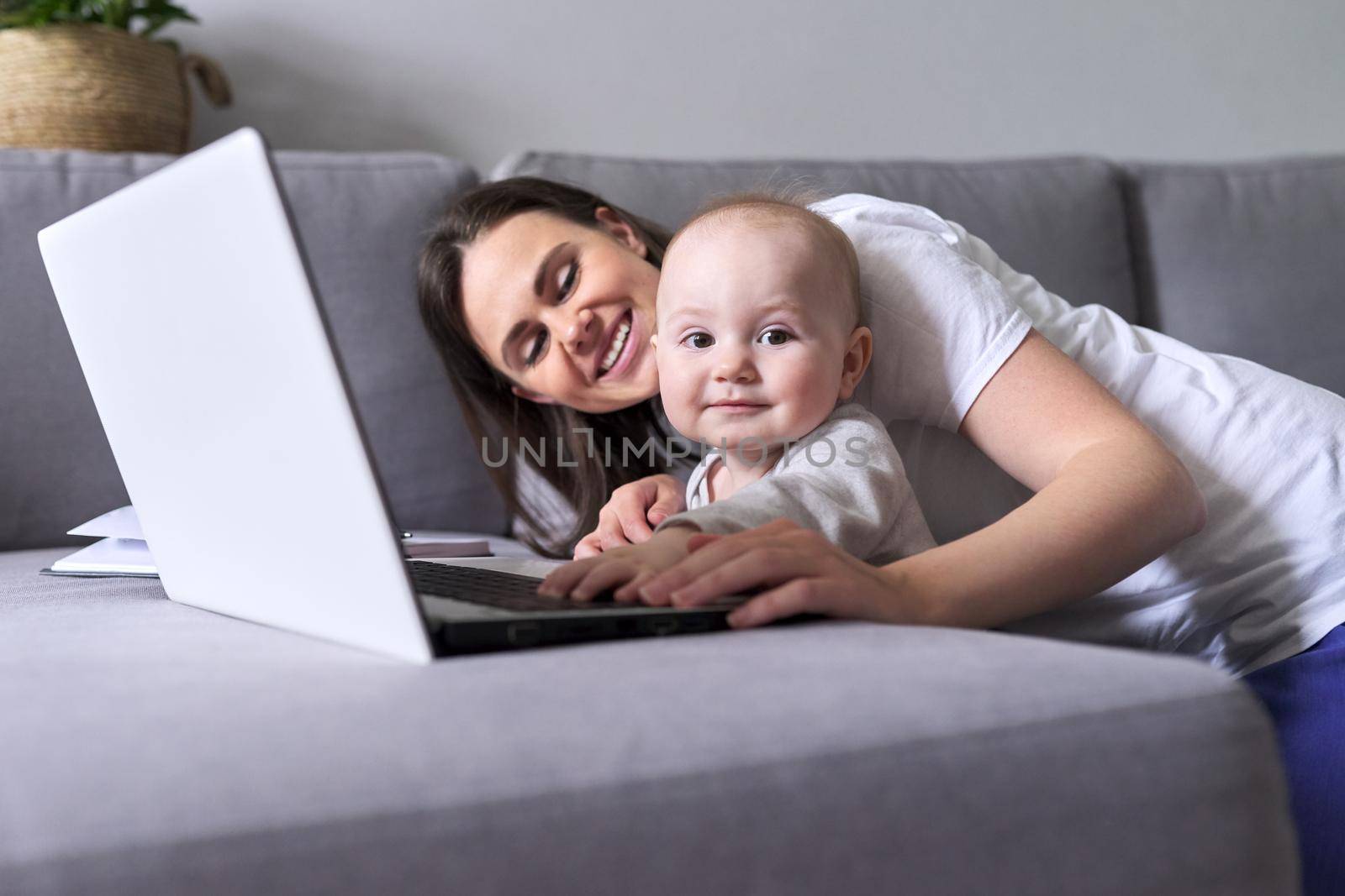 Young smiling mom with toddler baby look together at laptop monitor sitting at home on the couch