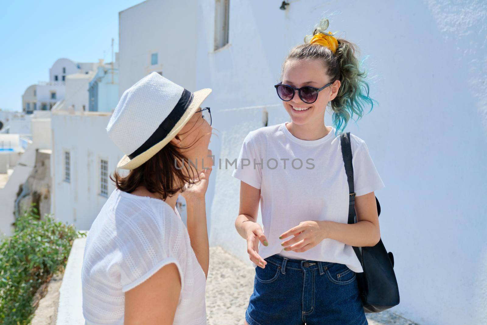Happy laughing talking mother and teenager daughter, white architecture tourist city background, sunny summer day