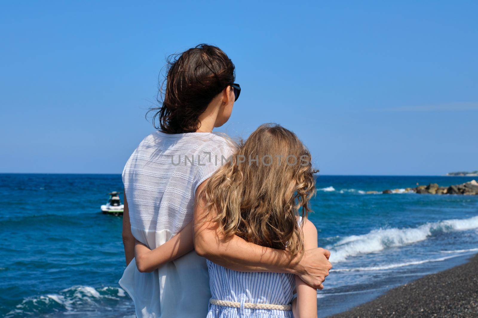 Happy mom and daughter kid hugging walking together along the beach, back view. Family, vacation, happiness, childhood, mother and child communication