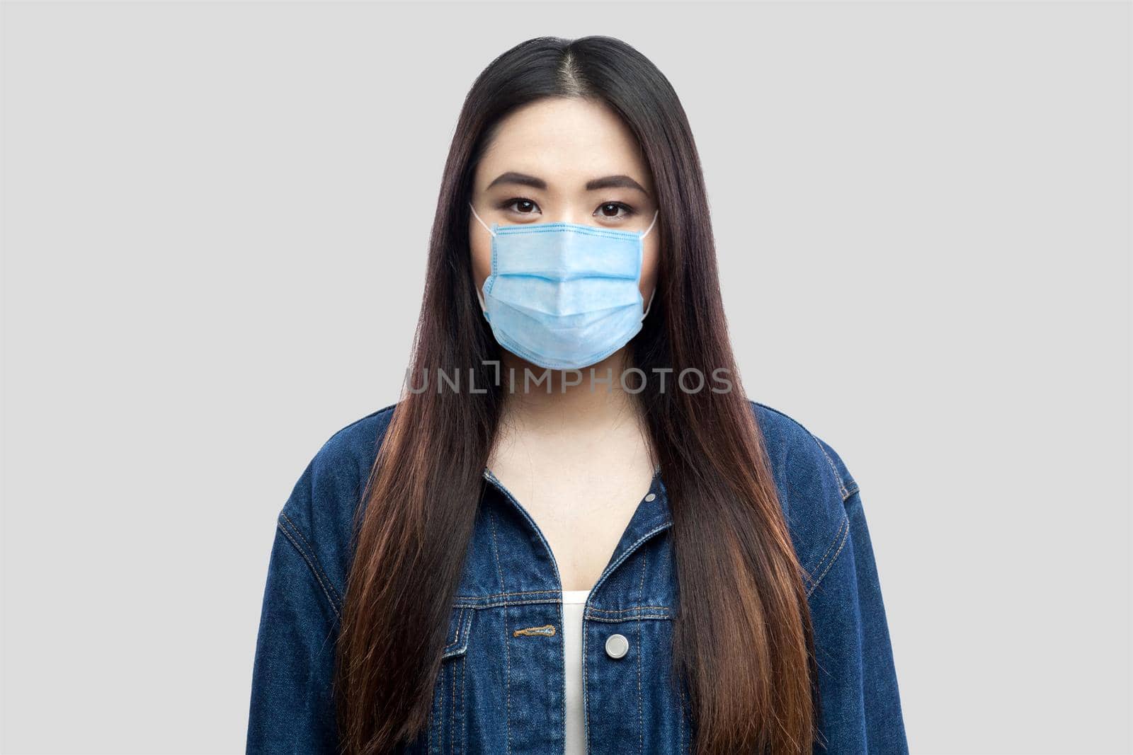 Portrait of beautiful brunette asian young woman with surgical medical mask in blue denim jacket standing and looking at camera. indoor studio shot, isolated on light grey background.