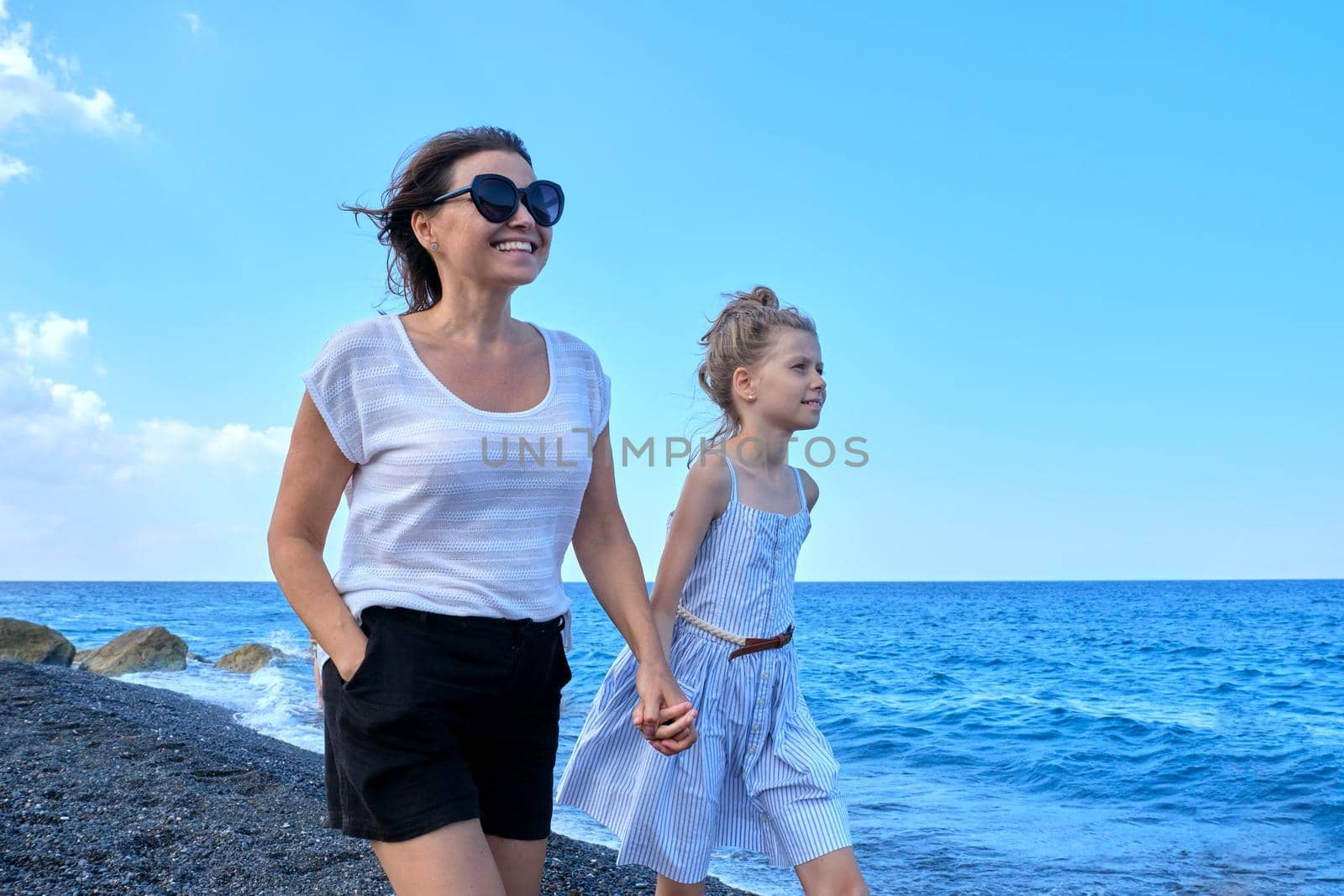 Happy mother and daughter kid walking together holding hands along the sea beach. Family weekend, travel, summer vacation concept