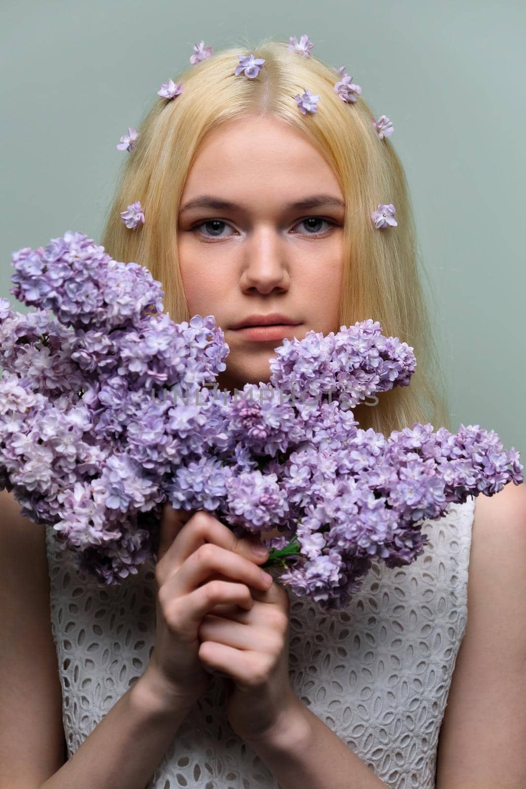 Portrait of young teen blonde girl with violet lilac flowers in hair and hands, pastel green studio background. Healthy skin and natural makeup for teenage girls