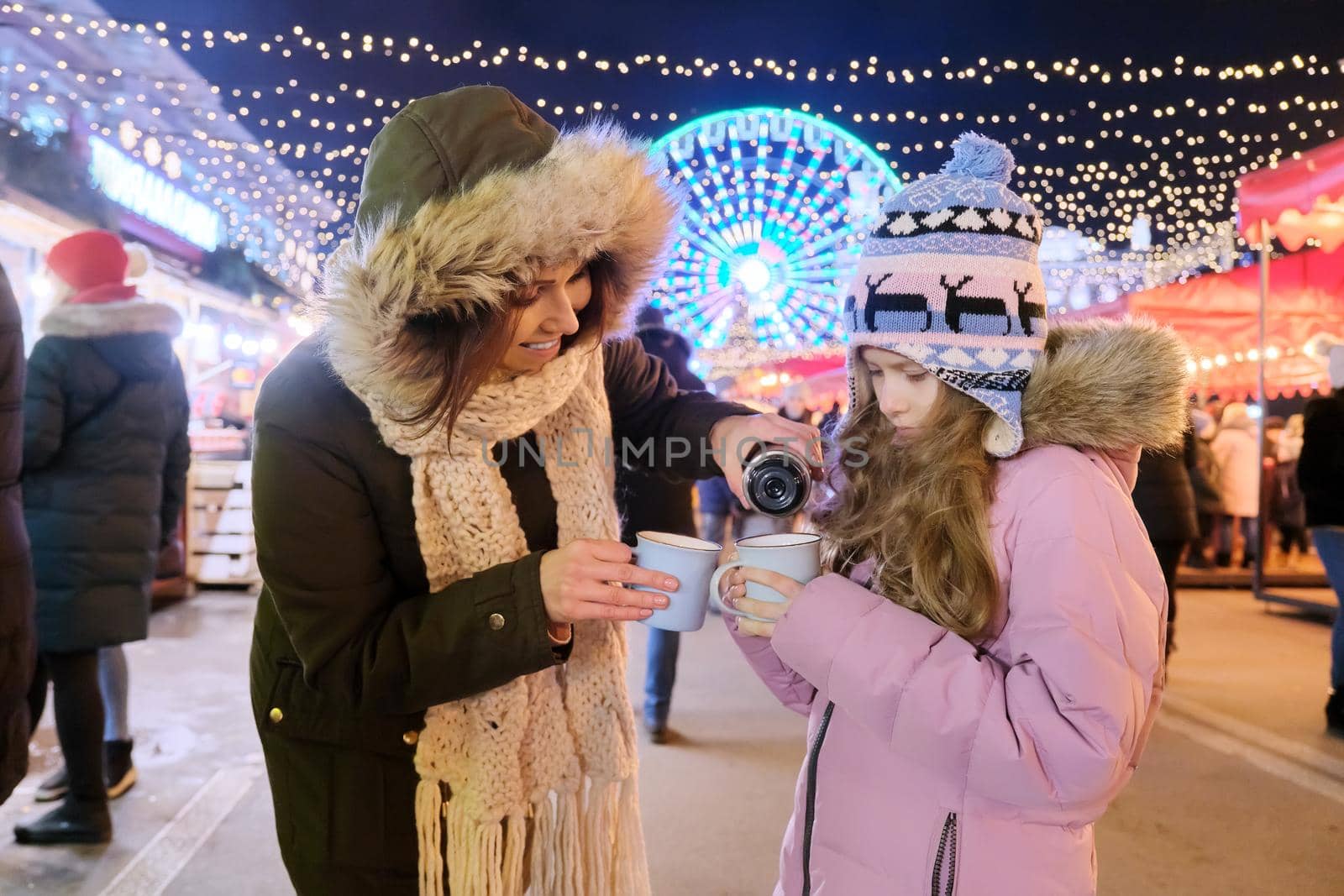Christmas and New Year holidays, happy mom and daughter kid walking together drinking hot mug tea at Christmas market, sparkling lights of garlands of evening city ferris wheel