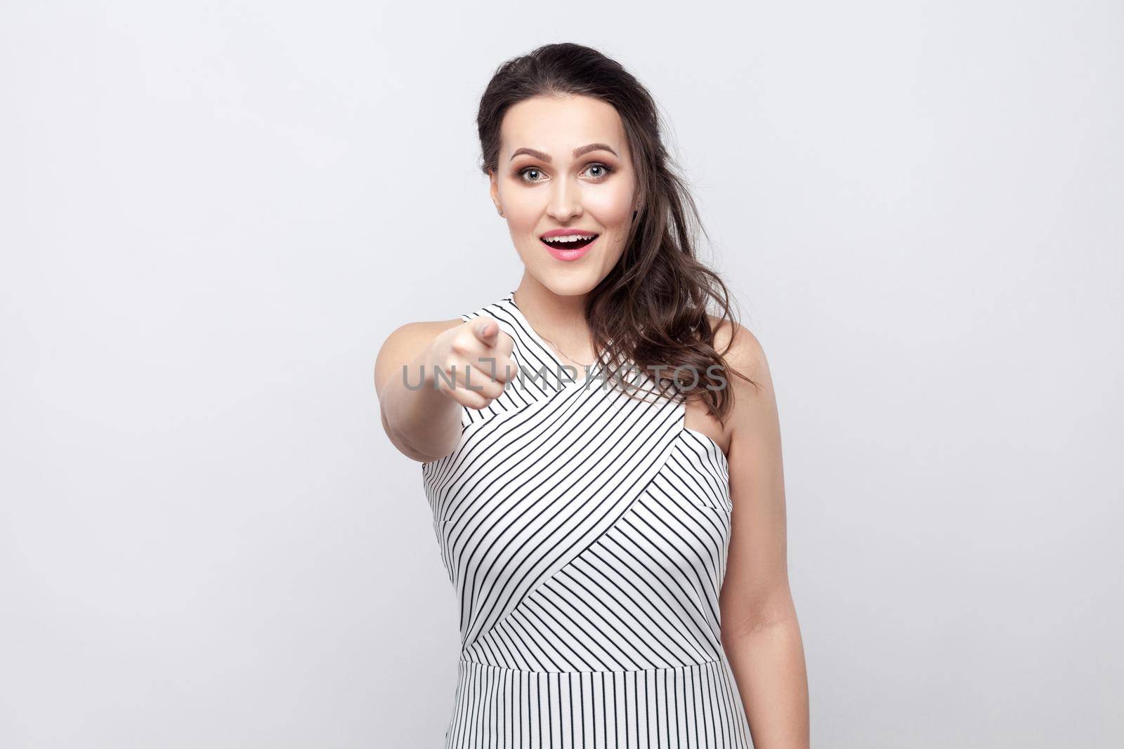 Portrait of amazed happy beautiful young brunette woman with makeup and striped dress standing, looking and pointing at camera with surprised face. indoor studio shot, isolated on grey background.