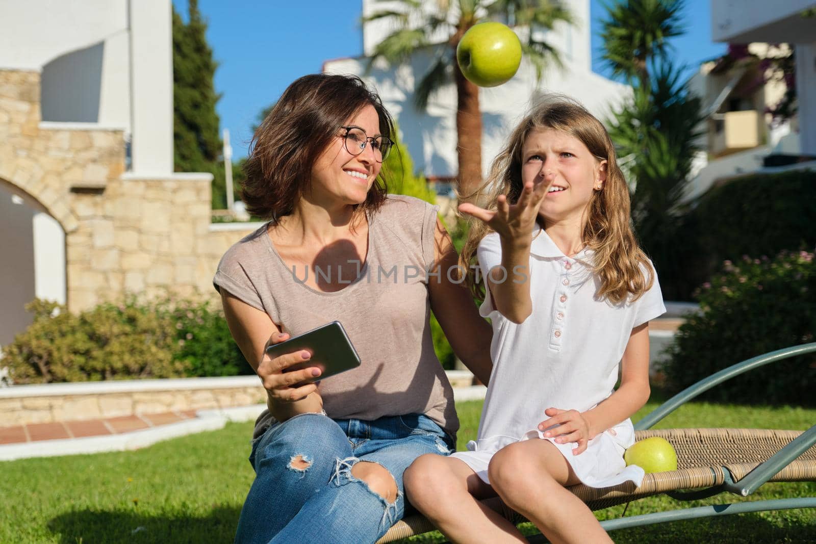 Mom and daughter child rest together sitting in an outdoor chair on the lawn, girl having fun with an apple