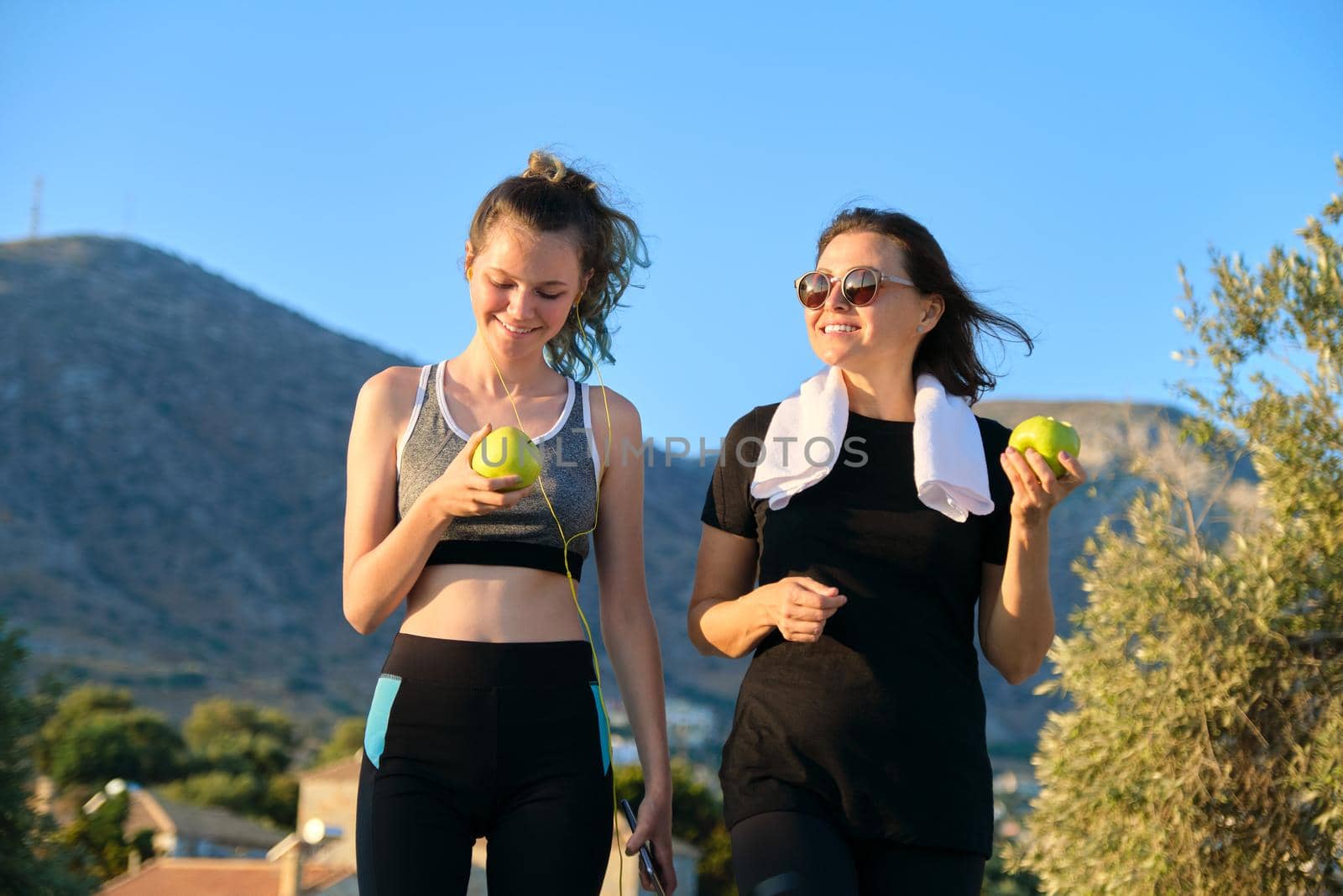 Active athletic healthy family lifestyle, smiling happy mother and teenage daughter eating apples, resting after jogging exercises. Beautiful sunset mountain nature background
