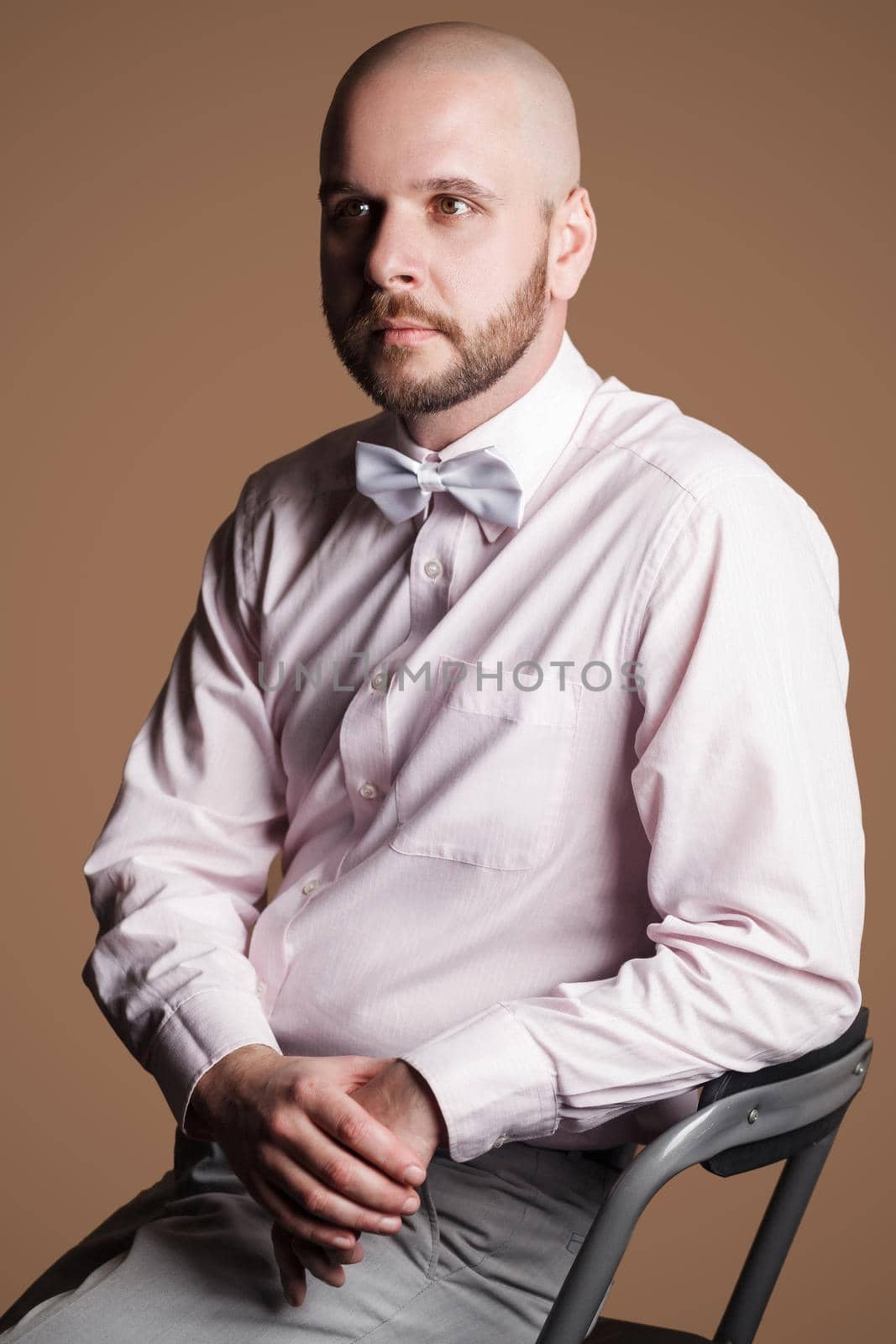 Profile side view portrait of handsome bearded bald man in light pink shirt and white bow, sitting on chair and looking away with thoughtful serious face. studio shot, isolated on brown background.