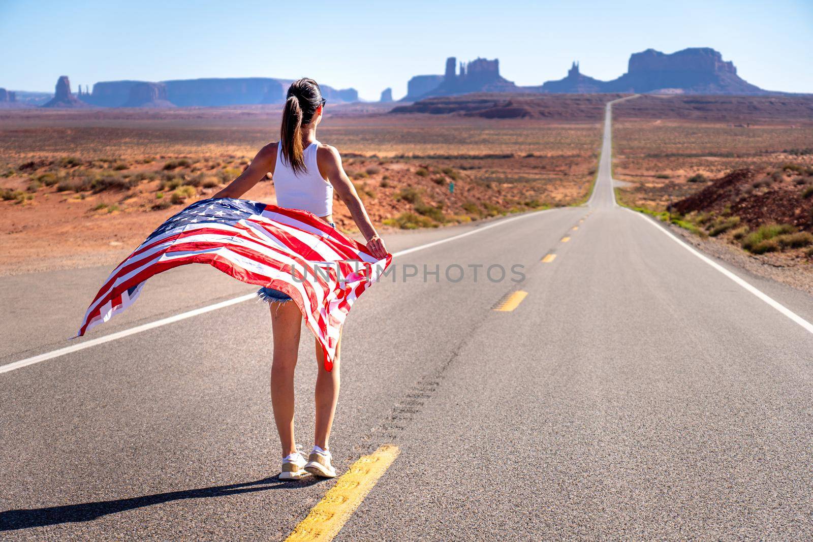 Unknown brunette girl from behind in Monument Valley holding a United States of America flag, wearing short jeans and a white tank top