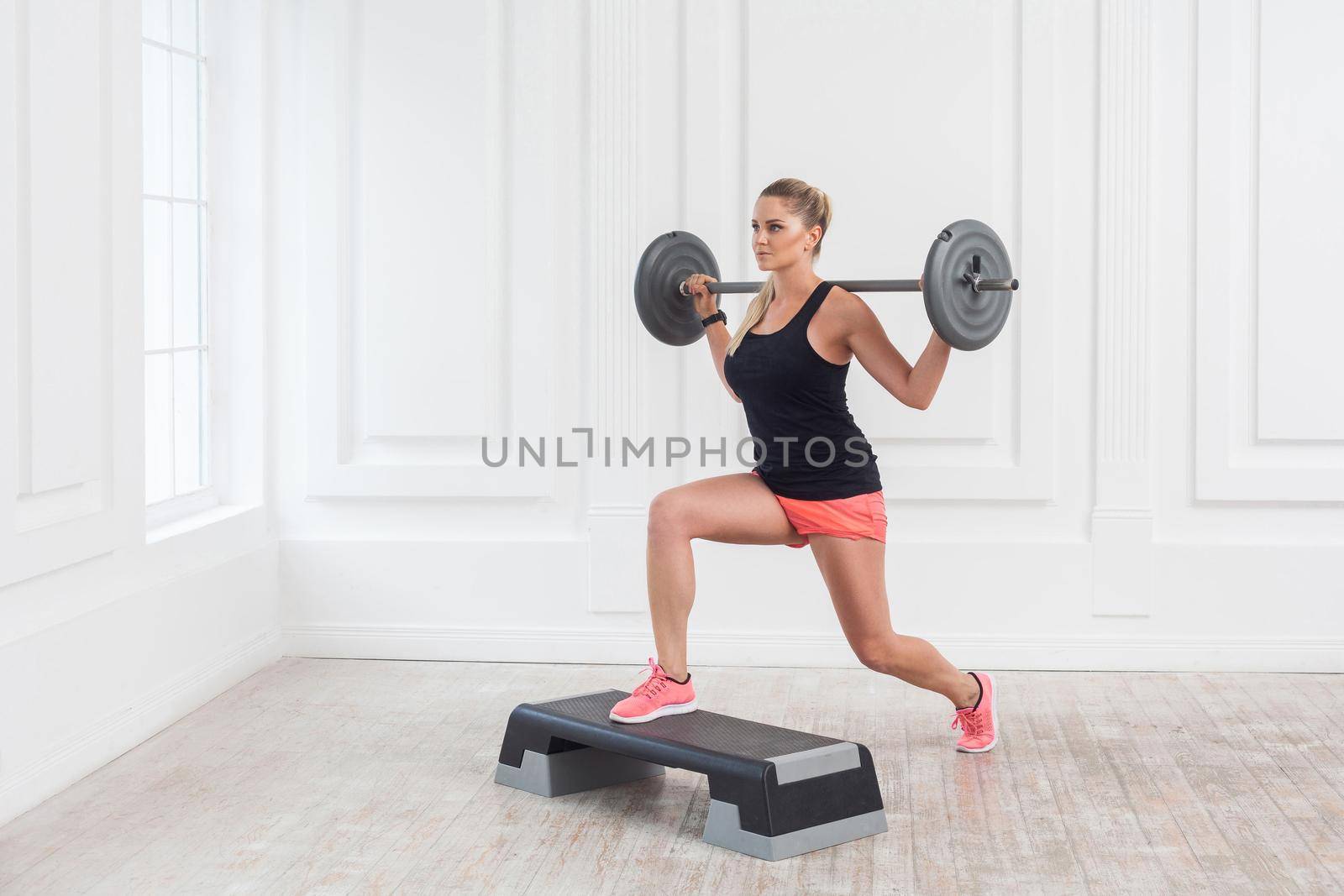 Portrait of young athletic beautiful bodybuilder woman in pink shorts and black top holding barbell on shoulders and doing squats with one leg on step at the gym on white wall. indoor, studio shot