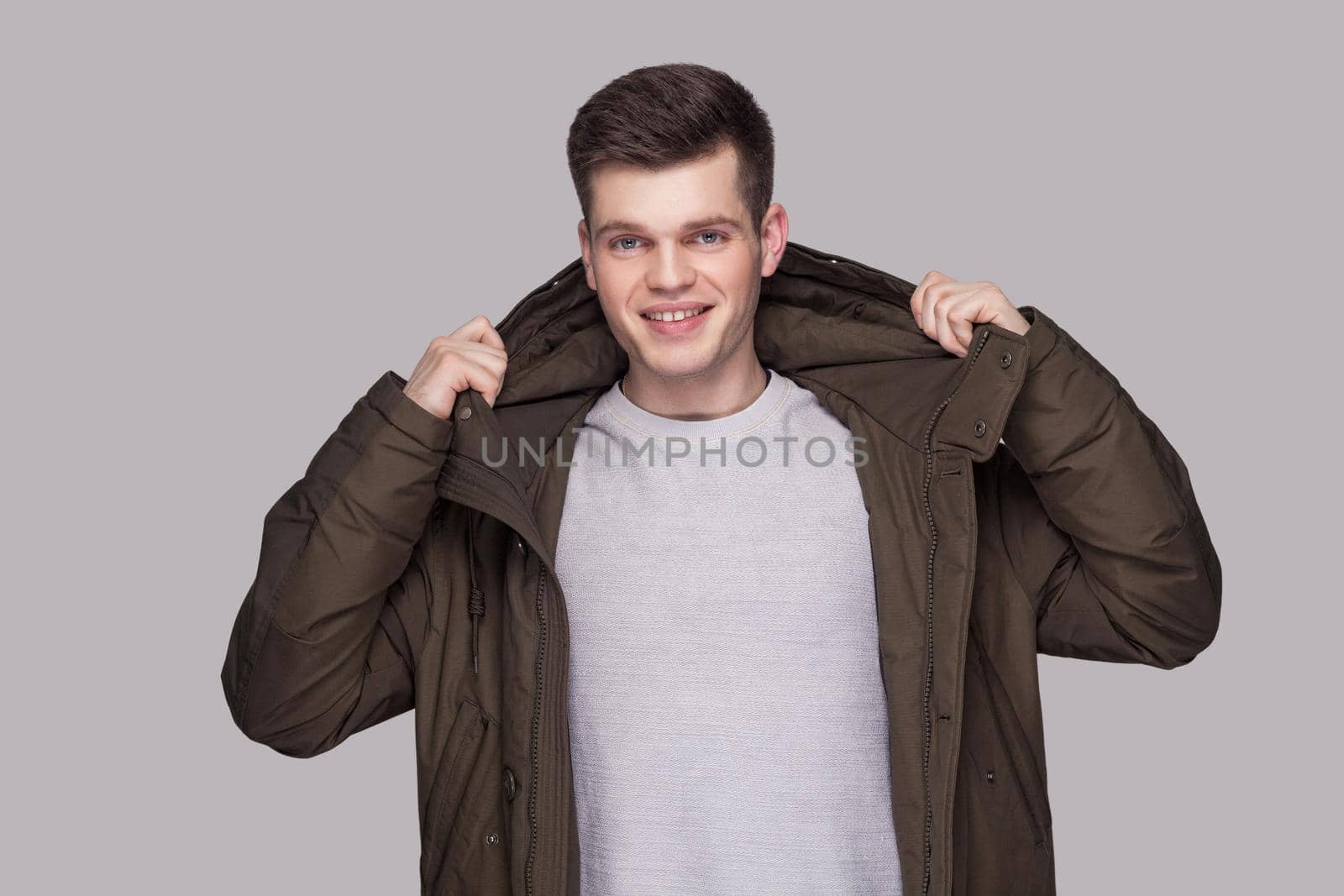 Portrait of young happy handsome man standing in light gray shirt and dark green parka looking at camera with toothy smile. indoor studio shot, isolated on gray background.