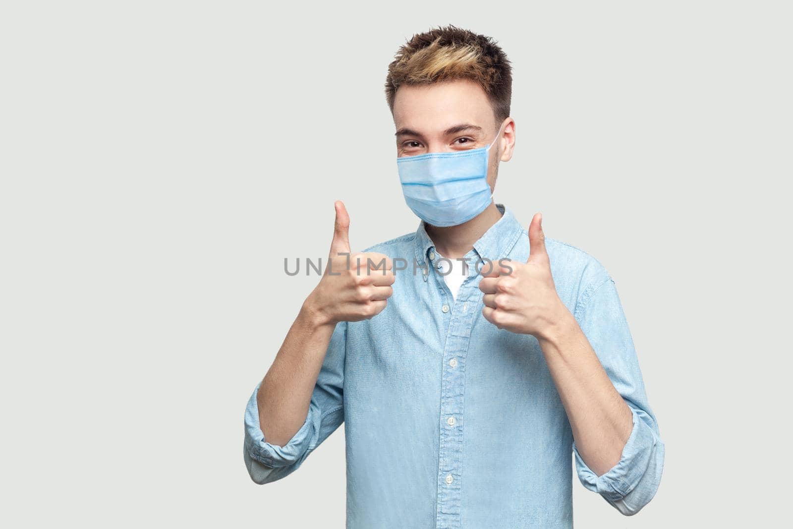 Portrait of happy satisfied successful handsome young man with surgical medical mask in light blue shirt standing, thumbs up and looking at camera. indoor studio shot on grey background copy space.