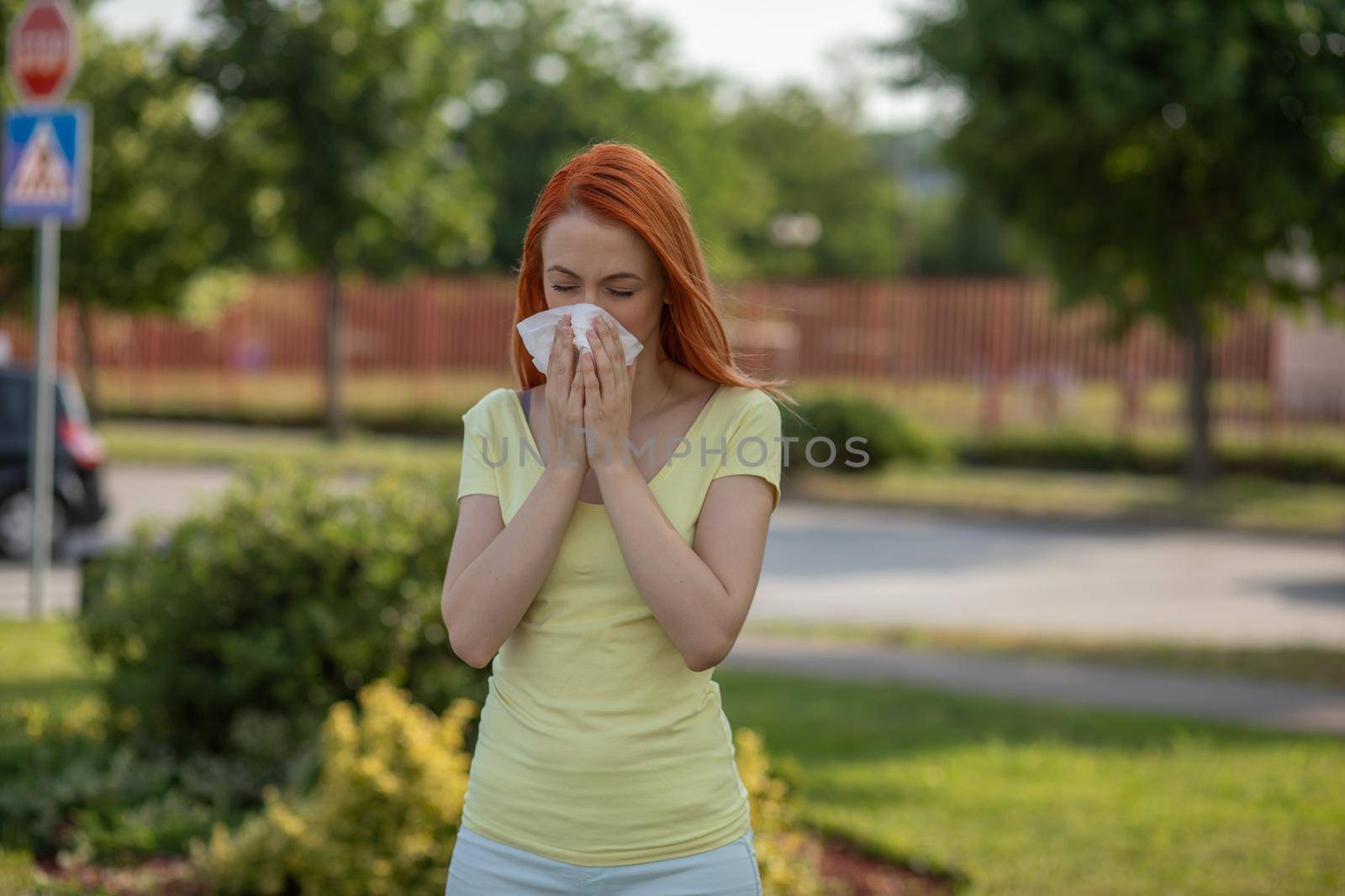 Young redhair woman sneezing in summer in front of blooming tree. Pollen Allergy symptoms