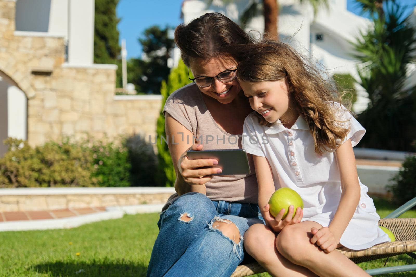 Mom and daughter kid using smartphone for video call. Family sitting on an outdoor chair on lawn in the yard
