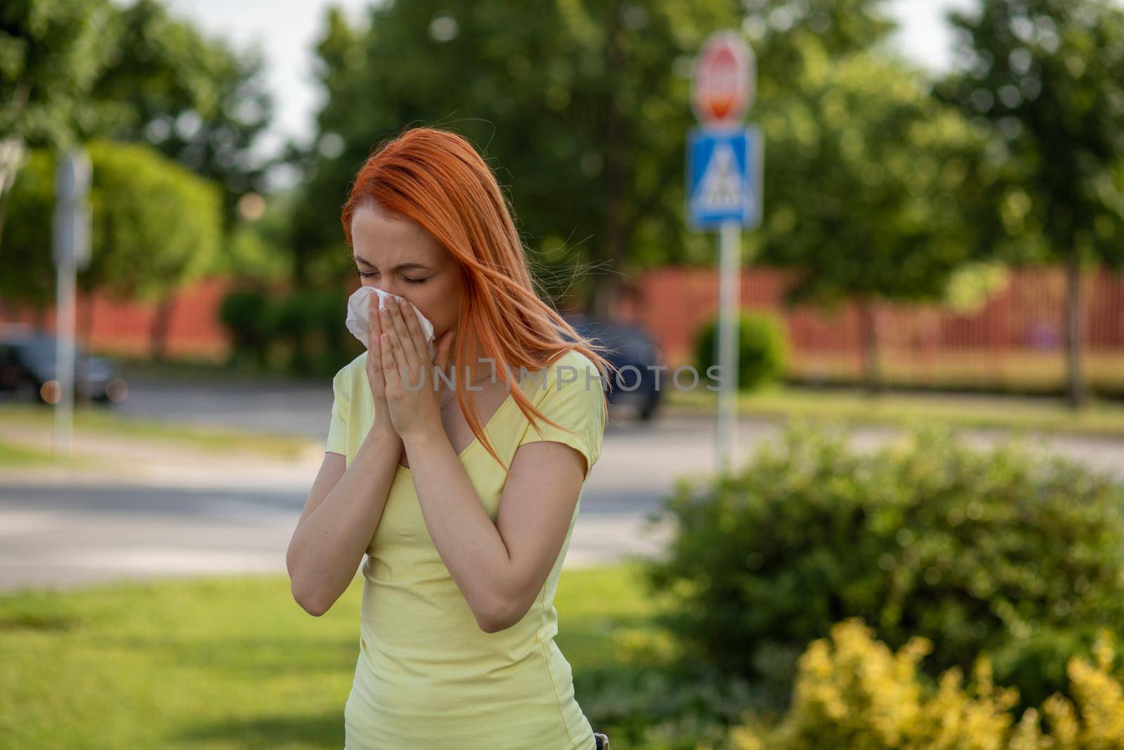Young redhair woman sneezing in summer in front of blooming tree. Pollen Allergy symptoms