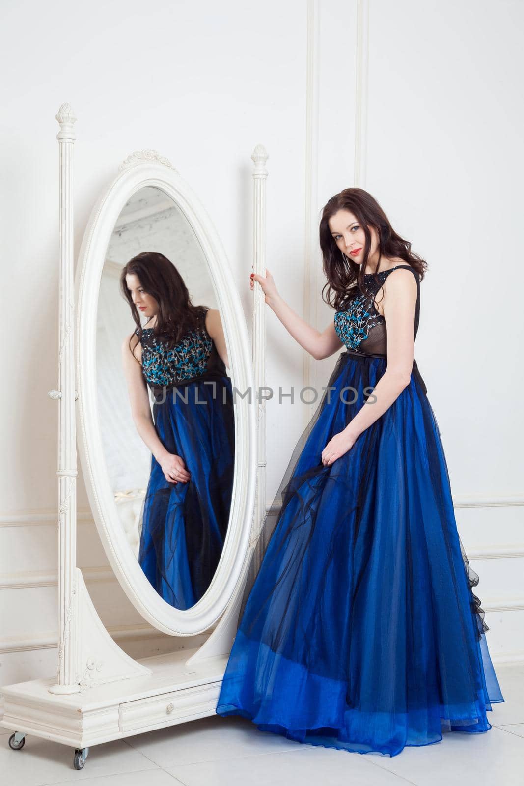 Gorgeous brunette woman in fashionable dress posing near big mirror at white room and looking at camera and smiling. indoor studio shot.