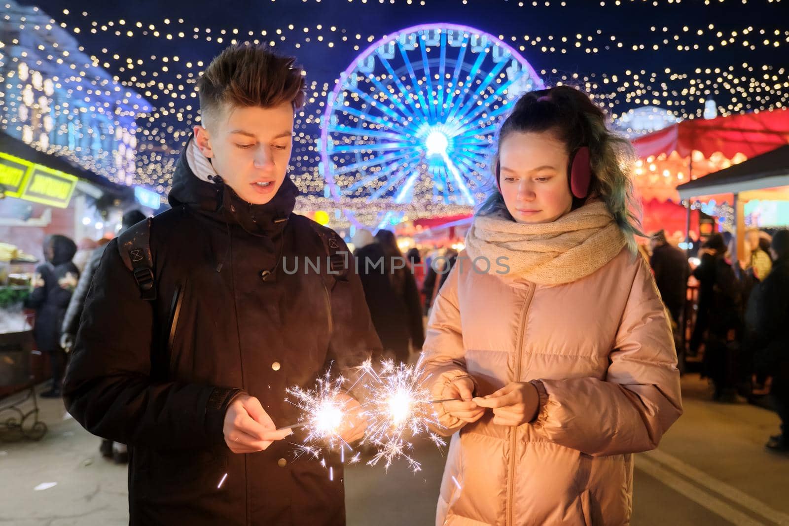 Christmas and New Year holidays, happy teenage couple with sparklers celebrating and having fun at Christmas market, sparkling lights of garlands of evening city ferris wheel background