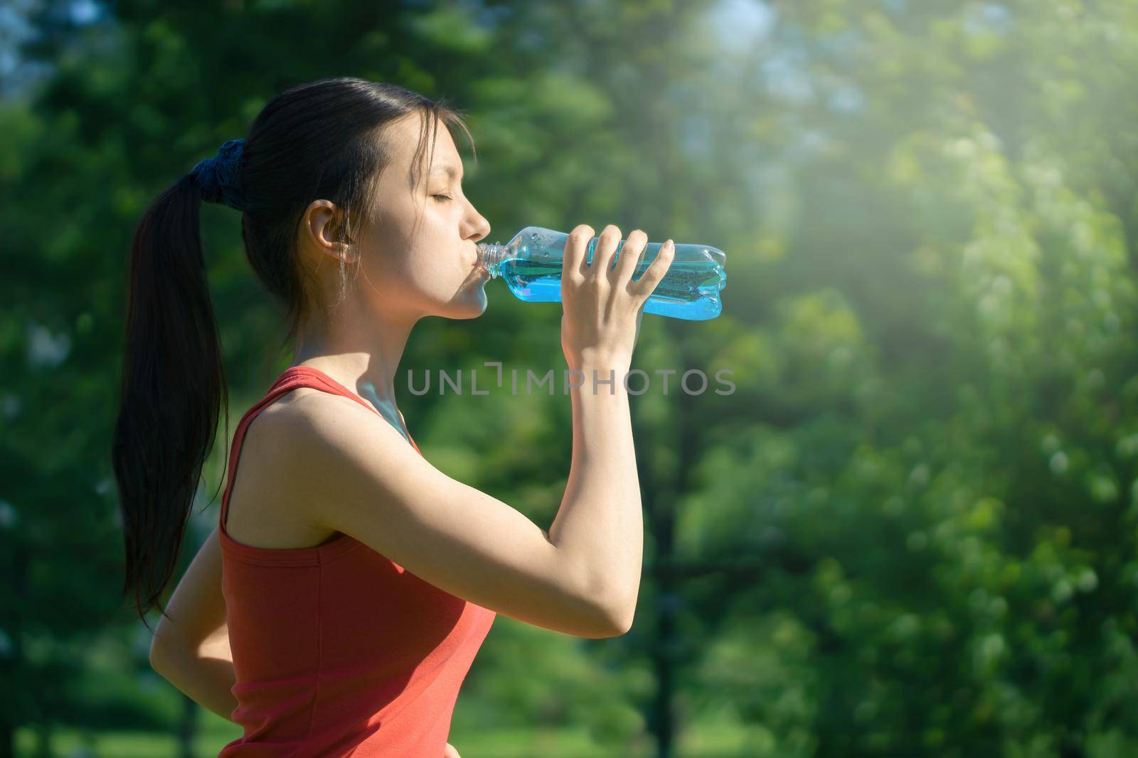 Beautiful young woman jogging in the morning in the park drinking blue isotonic from a bottle by galsand