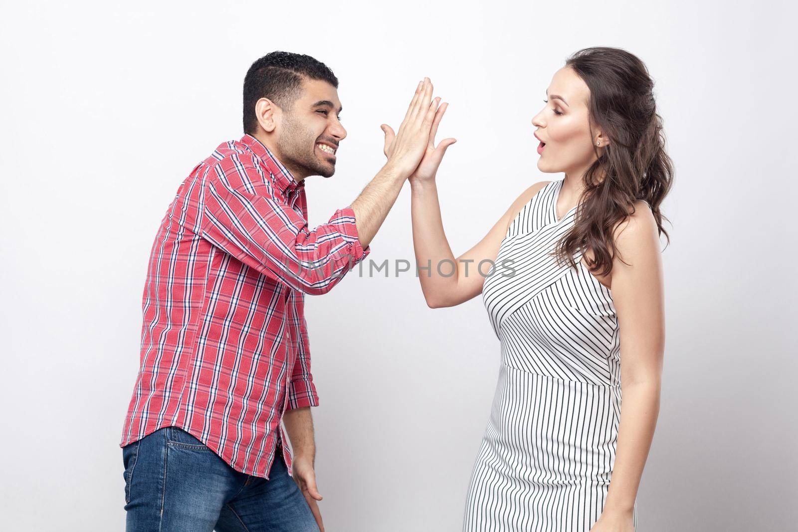 high five. Profile side view portrait of happy handsome man in red checkered shirt and beautiful woman in white striped dress standing and celebraiting victory. indoor studio shot on grey background.
