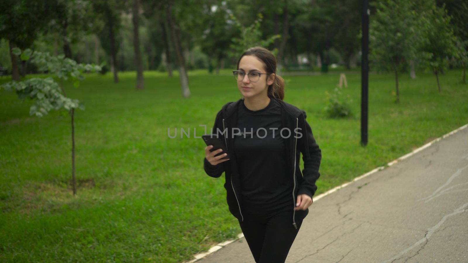 A young woman is jogging with a phone in her hand and rejoices. Asian girl with glasses is running with the application in the phone move to earn. 4k