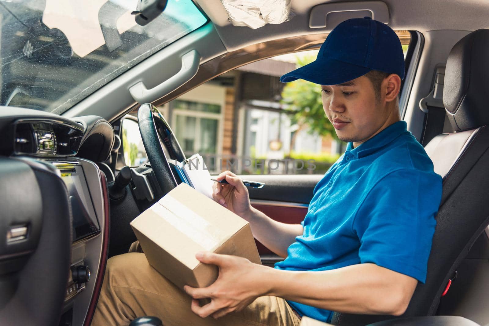 delivery man courier in uniform hold documents clipboard checking list parcel post boxes inside a car by Sorapop