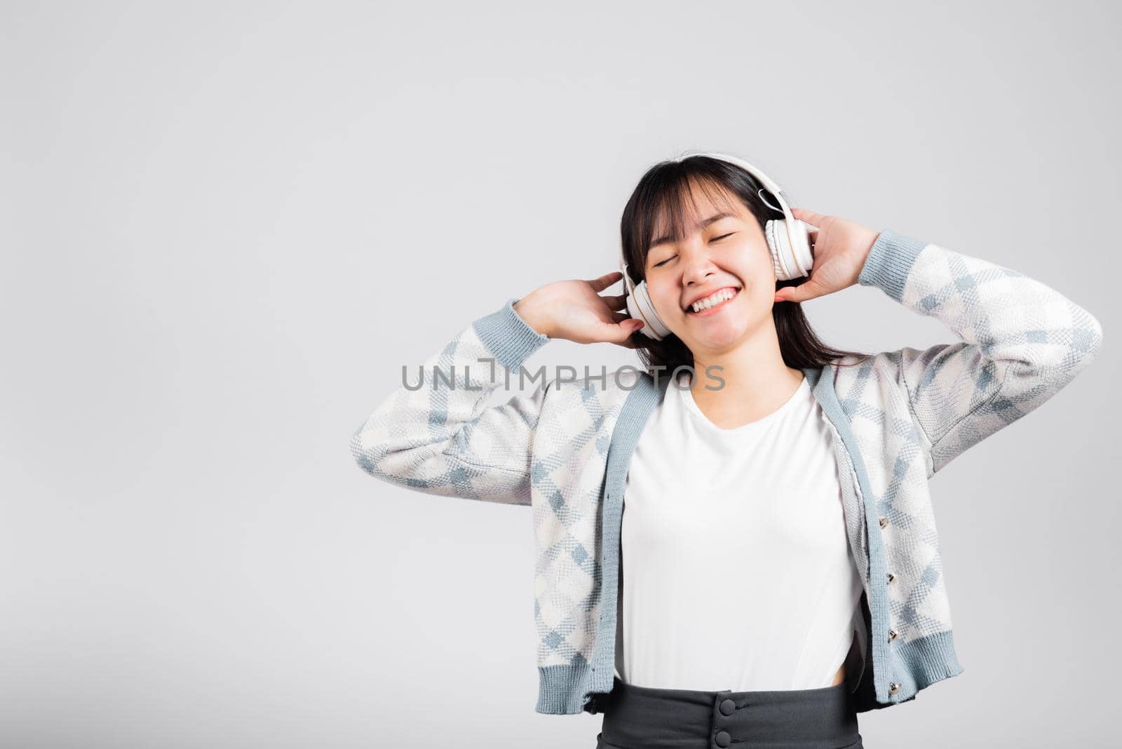 Woman excited smiling listening to music radio in bluetooth headphones and holding phone studio shot isolated white background, happy Asian young female listen modern technology mobile phone