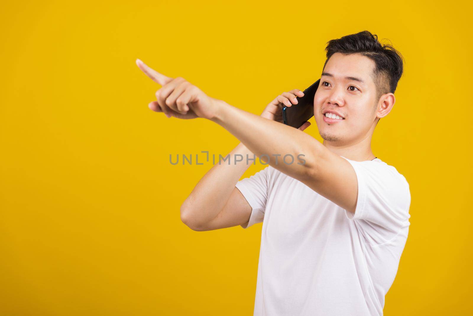 Asian handsome young man smiling positive showing smartphone blank screen and pointing finger to side away, studio shot isolated on yellow background, making successful expression gesture concept