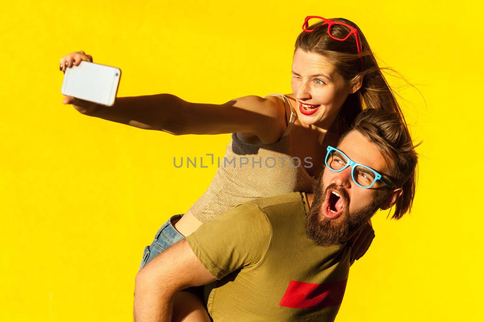 Young adult hipsters. Bearded man shout, freckled brunette woman wink, toothy smile and making selfie piggyback boy. Yellow background. Studio shot