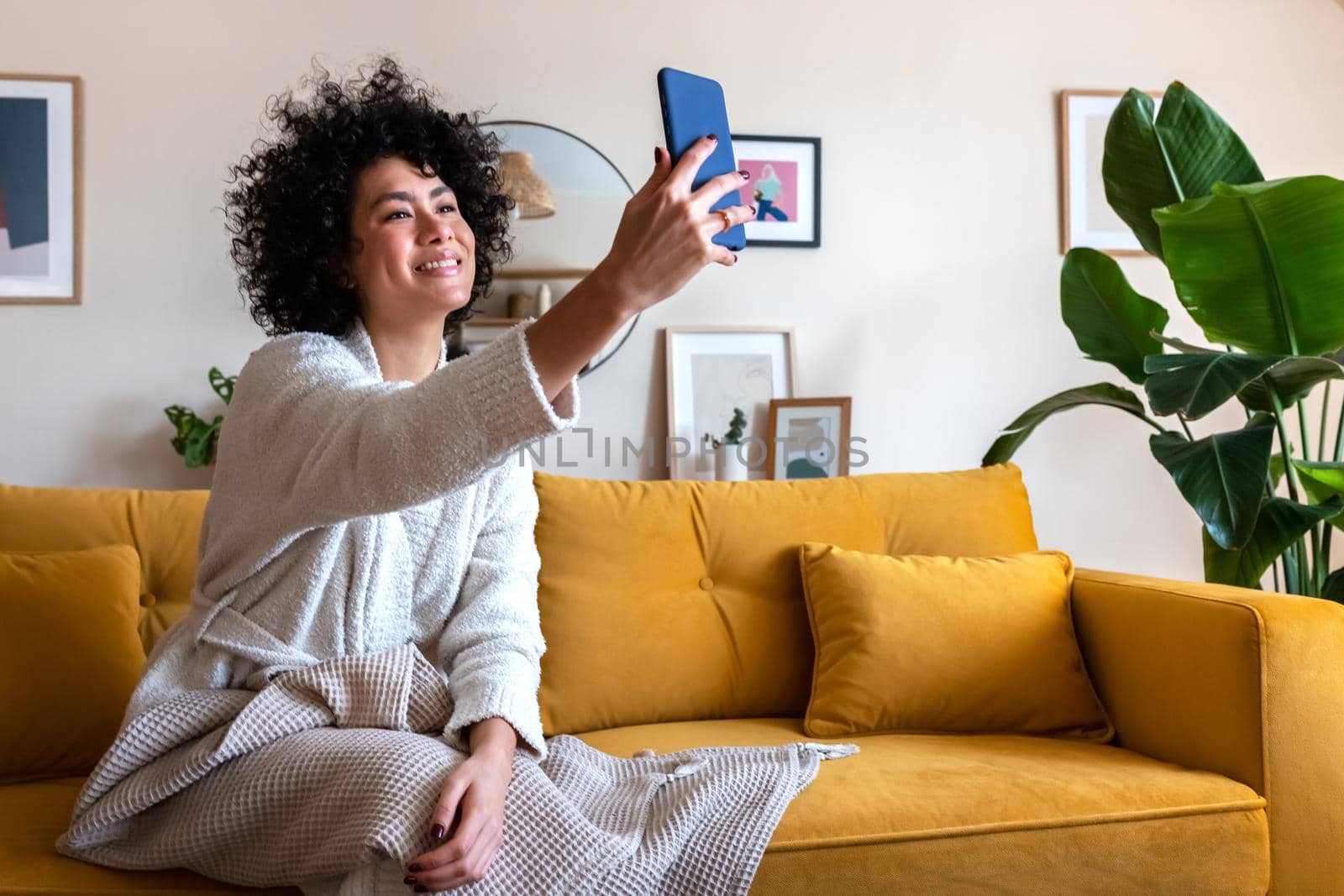 Smiling African american woman sitting on couch taking selfie photo at home using mobile phone. by Hoverstock